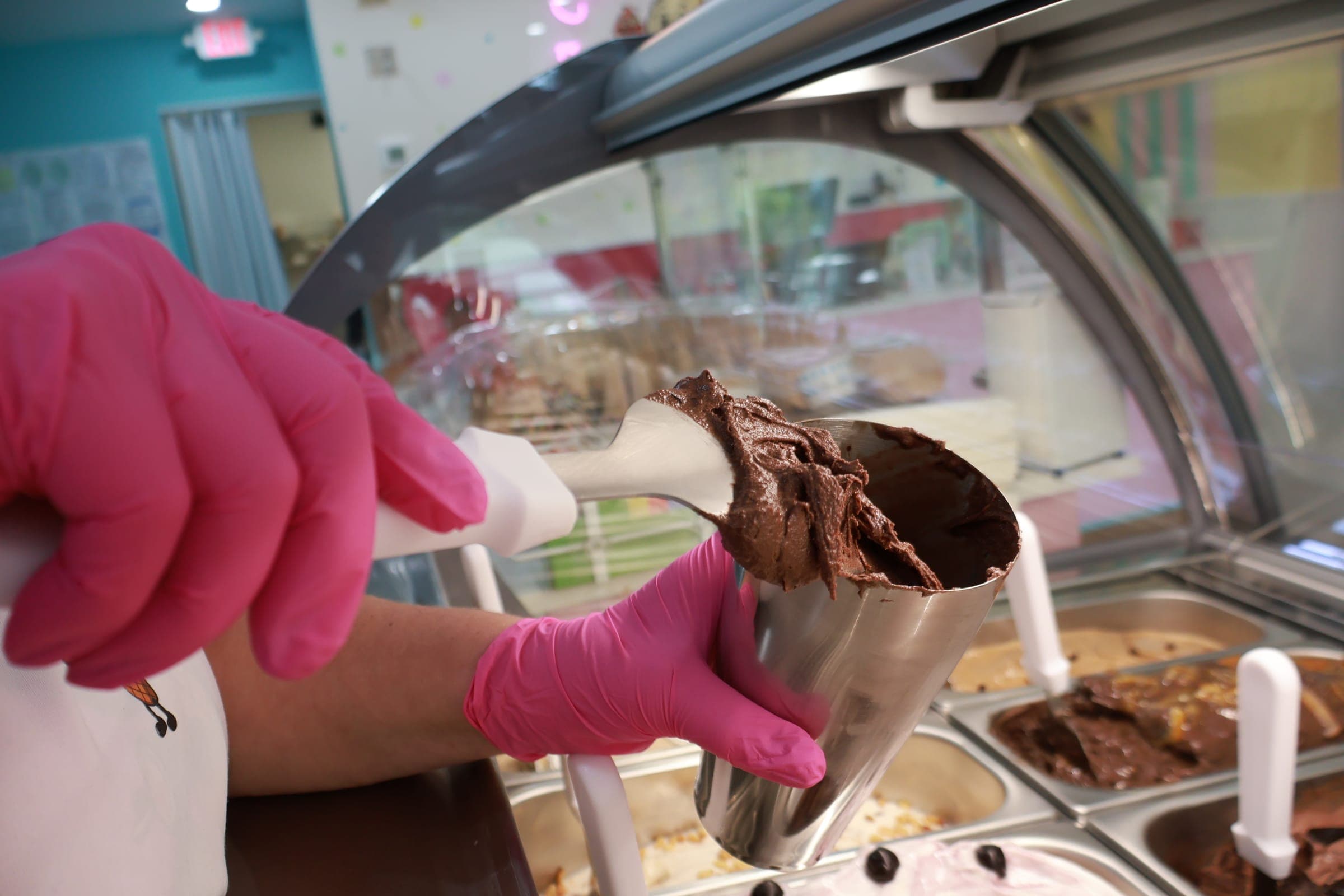 A gloved hand scooping chocolate gelato into a cup at the counter